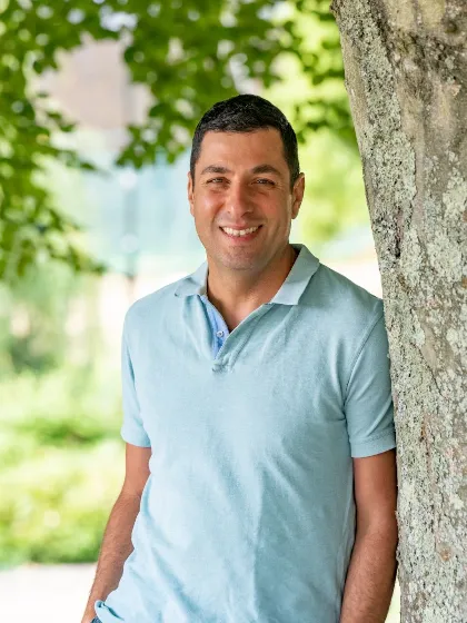 Professor Ali Tavassoli leaning against a tree in pale blue polo shirt