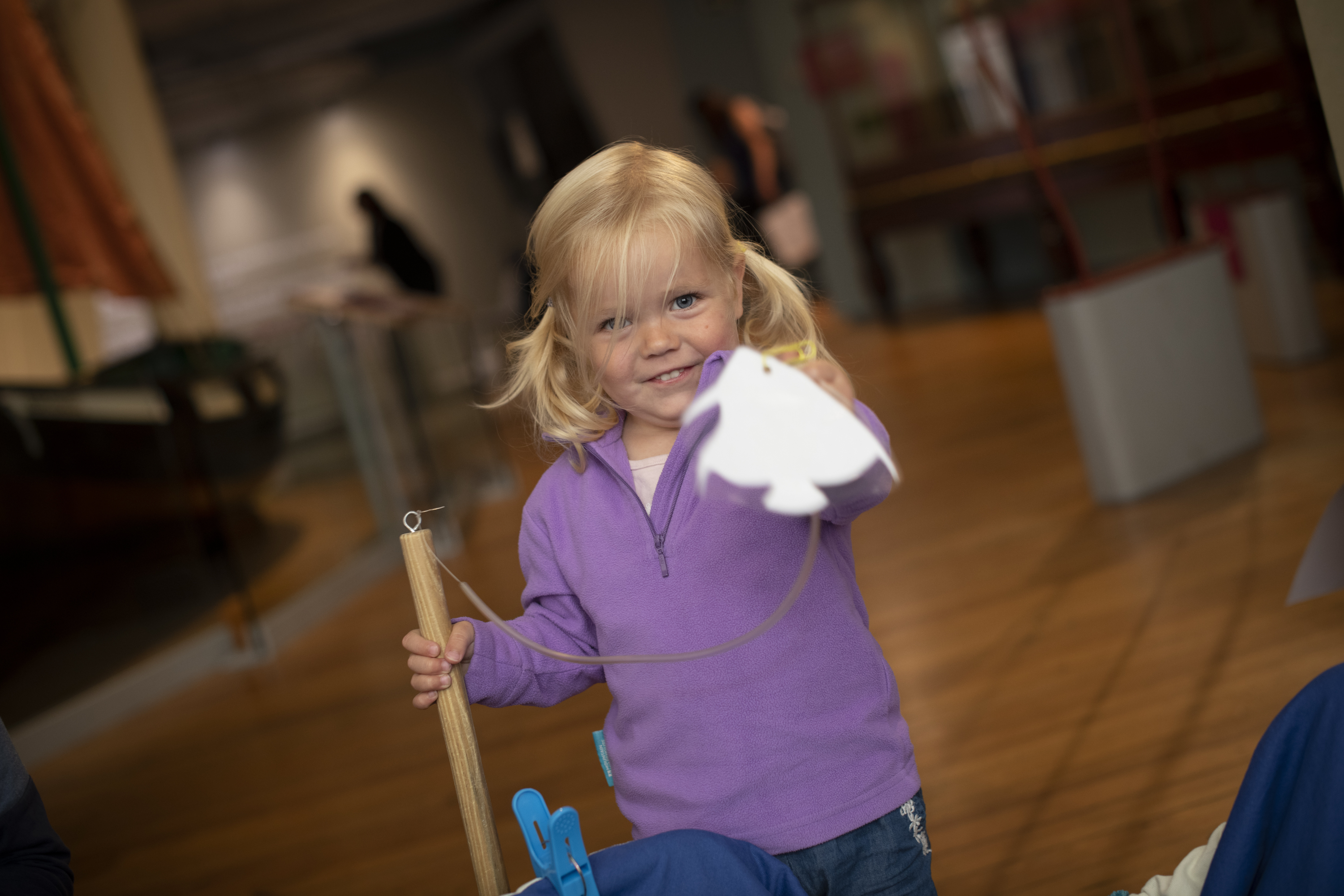A little girl in a purple jumper points at the camera while holding something in her left hand and gripping a wooden post with her right. A little girl in a purple jumper points at the camera while holding something in her left hand and gripping a wooden post with her right.