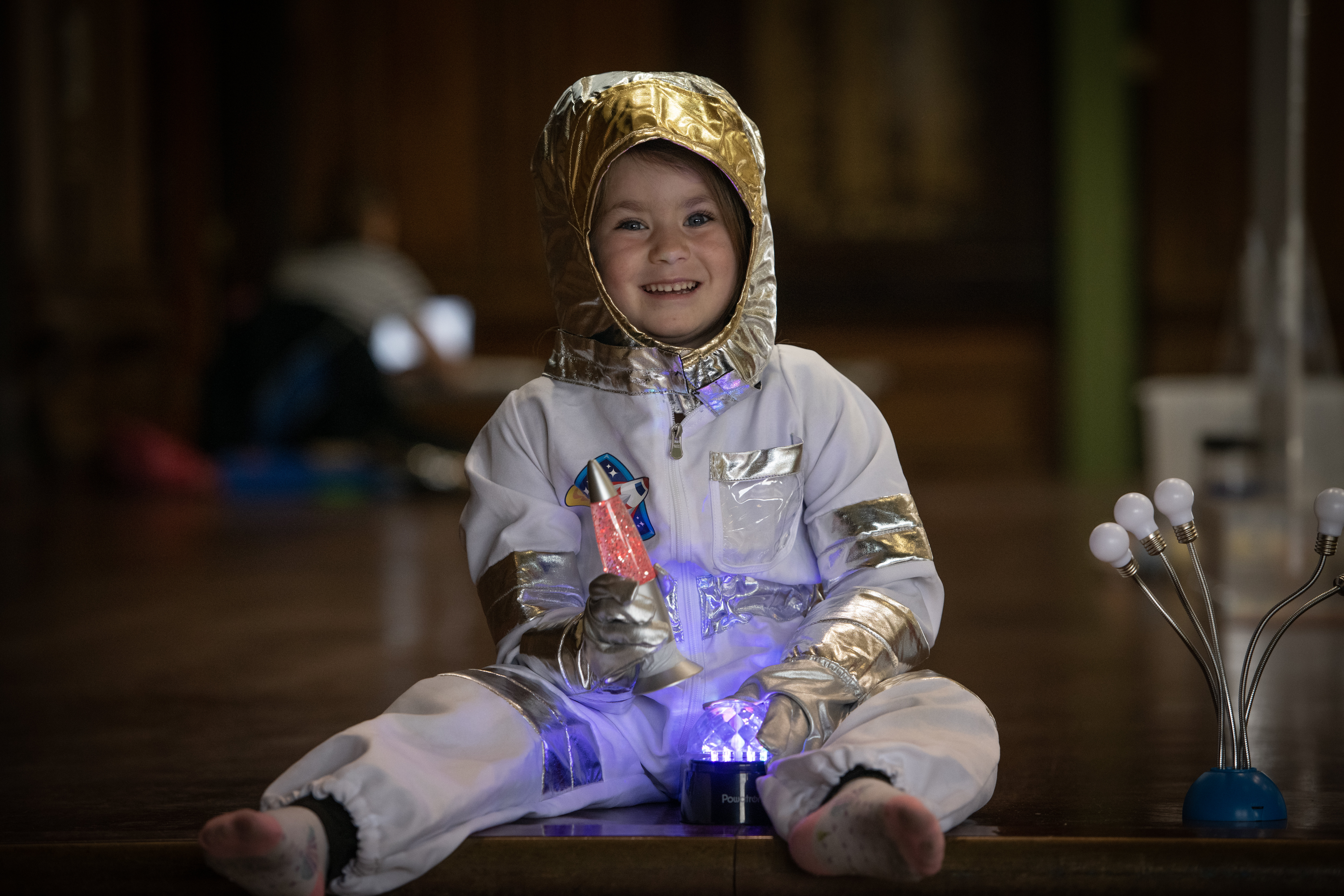 A little girl in an astronaut costume smiles as she holds a lava lamp and sits in front of a little light A little girl in an astronaut costume smiles as she holds a lava lamp and sits in front of a little light